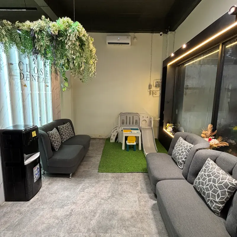 Ruang tunggu Close Dental dengan tanaman gantung, sofa, dan area bermain anak